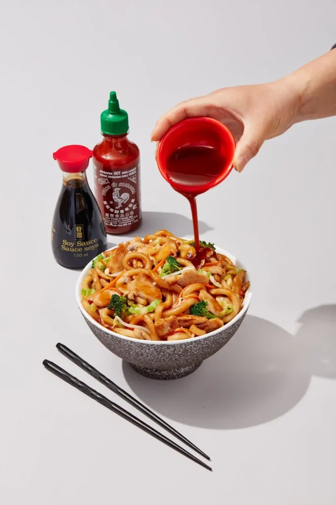 Bowl of udon noodles with broccoli and chicken, with sriracha being poured, alongside soy sauce and chopsticks.