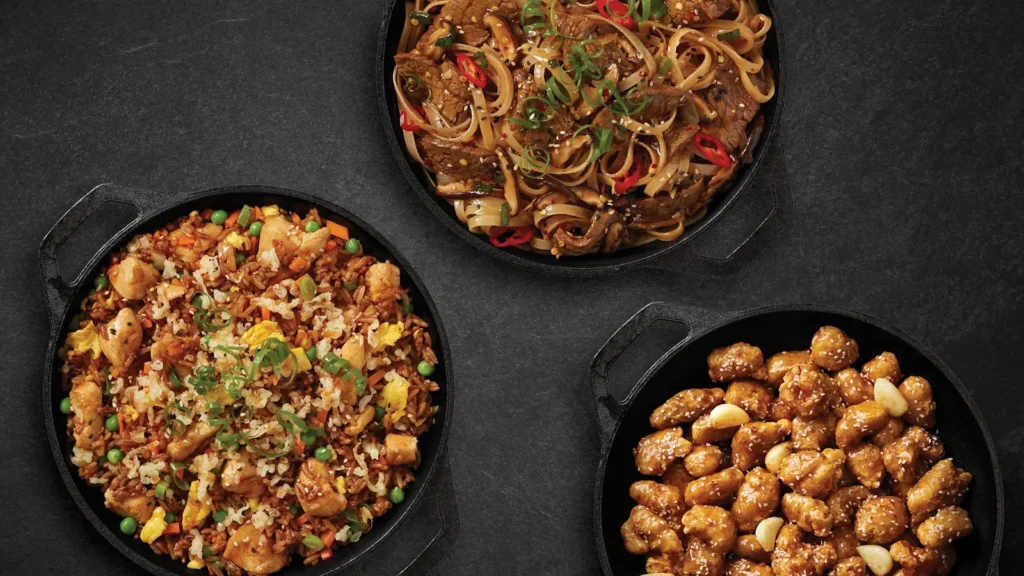 Three cast iron skillets with chicken fried rice, spicy beef noodles, and sesame chicken on a dark surface.