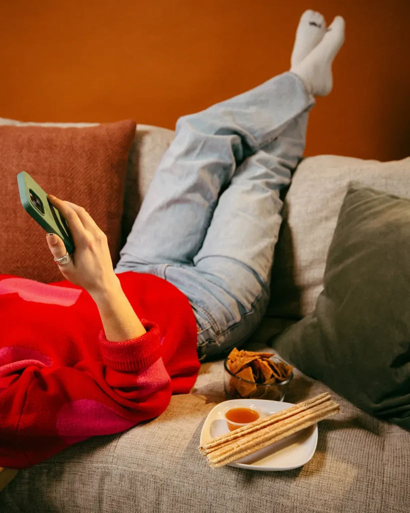 Person relaxing on a couch with their phone, churros with caramel sauce, and tortilla chips nearby.