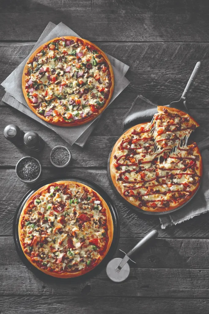 Three freshly baked pizzas on a dark wooden table: veggie, meat lovers, and BBQ chicken, with a pizza cutter and seasonings.