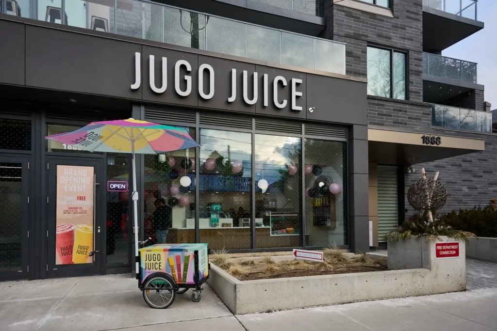 Jugo Juice storefront at street level of a modern condo building, with a grand opening sign, colourful umbrella and branded smoothie cart on the sidewalk.